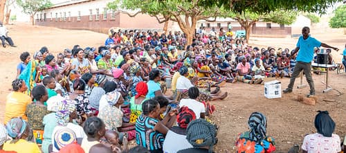 People sitting on the ground, listening to a man.